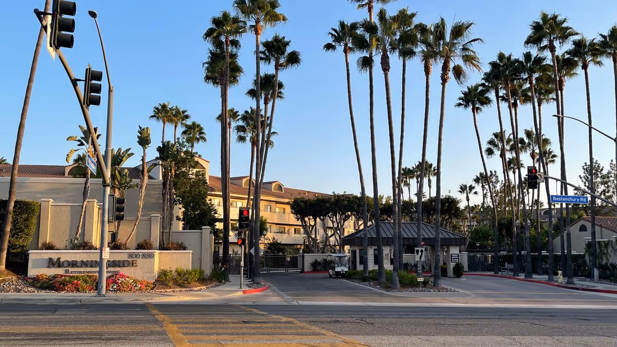 Entrance and front exterior of Morningside of Fullerton with tall palm trees, a stone sign, gated driveway, and traffic lights.