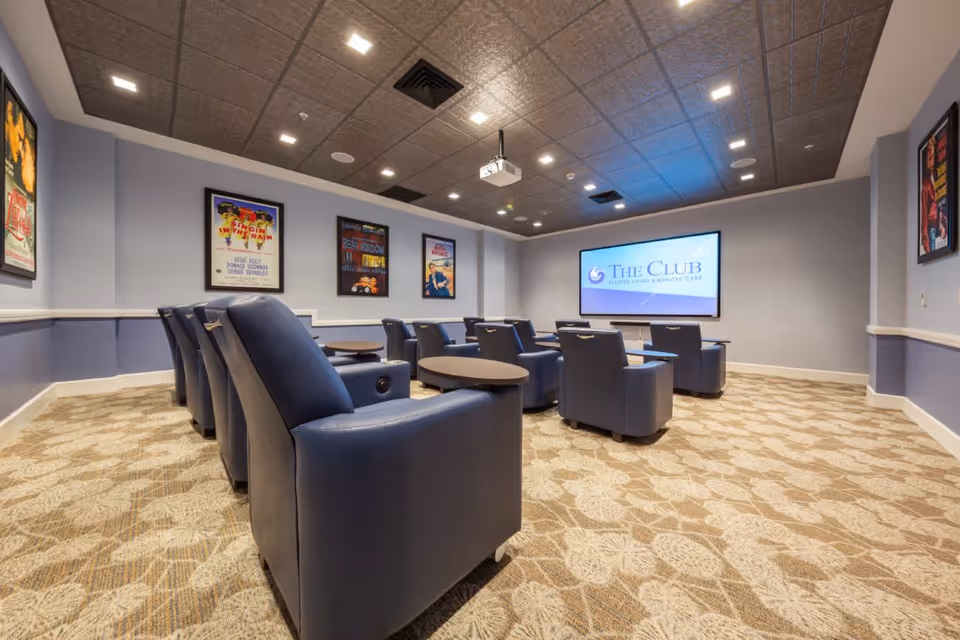 A cozy media room with several rows of blue recliner chairs facing a large screen displaying 'The Club Assisted Living & Memory Care'. The room has patterned carpet, light blue walls with white trim, movie posters framed on the walls, and a ceiling-mounted projector.