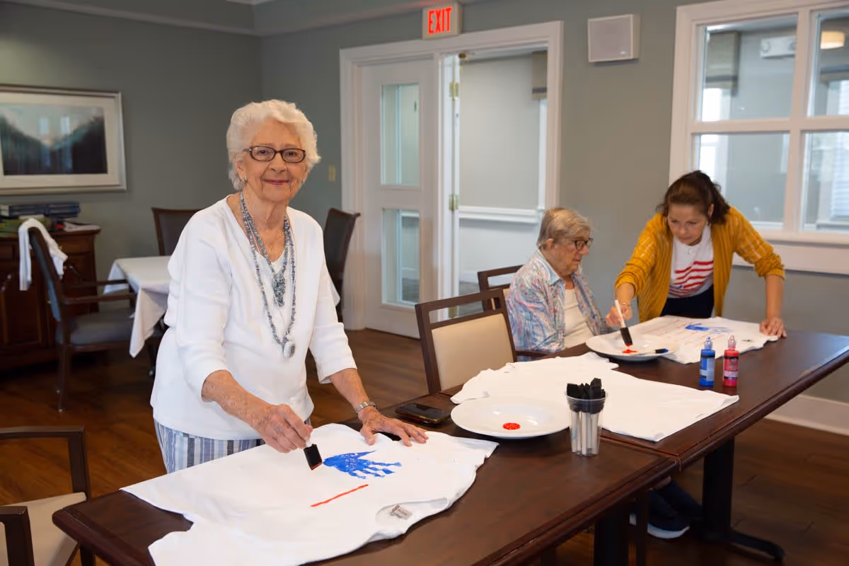An elderly woman with white hair and glasses stands at a table painting a blue handprint on a white t-shirt. Another elderly woman and a younger woman are seated at the same table, also painting on white t-shirts. The room has wooden floors, light gray walls, and large windows letting in natural light.
