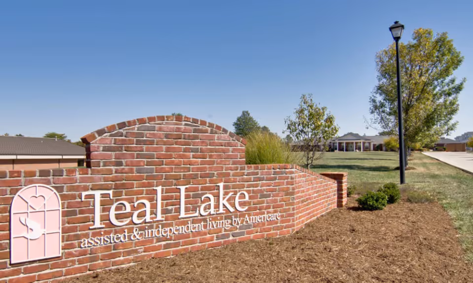 A brick sign for Teal Lake assisted and independent living by Americare is displayed outdoors on a landscaped area with mulch, small bushes, and trees. In the background, there is a building and a lamppost under a clear blue sky.