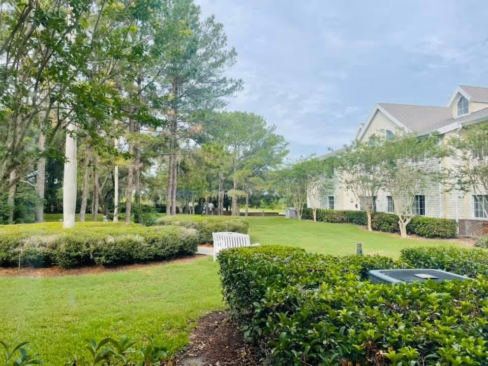 A landscaped outdoor area with green grass, trimmed bushes, and tall trees. A white bench is placed on a paved path near the center. To the right, there is a light-colored building with multiple windows and a sloped roof. The sky is partly cloudy.