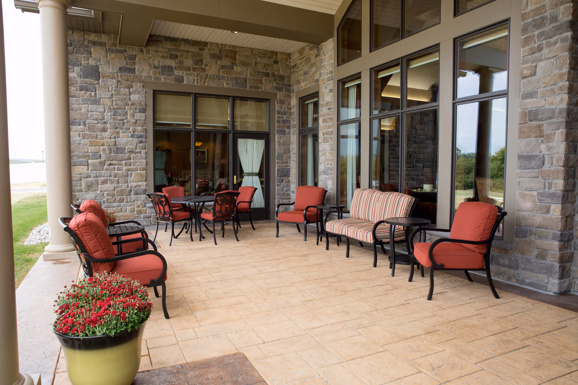 Covered outdoor patio area with stone walls and large windows. The patio is furnished with cushioned chairs, a striped cushioned bench, and a round table with chairs. A large planter with red flowers is visible in the foreground.