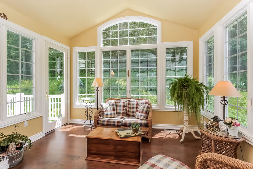 Bright sunroom with a plaid-cushioned wicker sofa, large arched windows, potted plants, and a wooden coffee table.