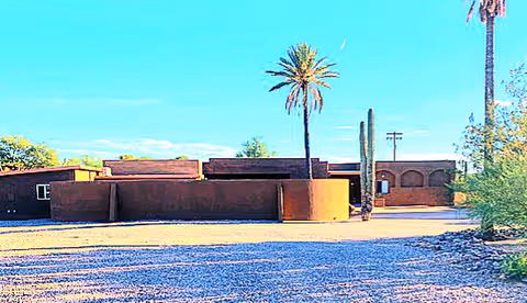 Single-story adobe-style building with palm trees and a tall cactus in a gravel desert yard under a bright blue sky.