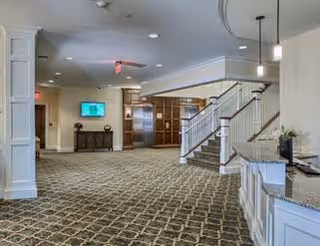 Spacious interior lobby area with patterned carpet, a staircase with white railings, a reception desk with a flower arrangement, and a TV mounted on the wall above a console table.