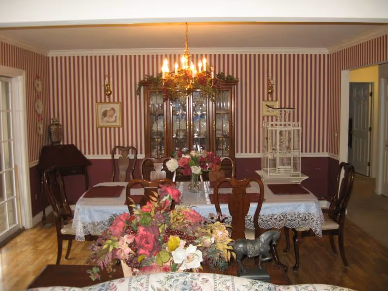 A traditional dining room with a wooden dining table covered with a lace tablecloth and set with floral centerpieces. The room features striped wallpaper in red and white, a wooden china cabinet filled with dishes, a chandelier with lit candles hanging above the table, and a decorative birdcage on a side table. There are wooden chairs around the table and hardwood flooring.