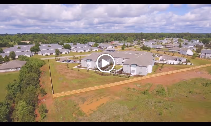 Aerial view of The Canopy at Warner Robins senior living facility showing multiple buildings with gray roofs surrounded by green trees and open land under a partly cloudy sky.