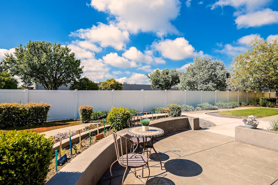 Outdoor patio area with a small round table and two metal chairs on a concrete surface. The patio is surrounded by low concrete walls and various green bushes and plants. A white fence and several trees are visible in the background under a blue sky with scattered clouds.