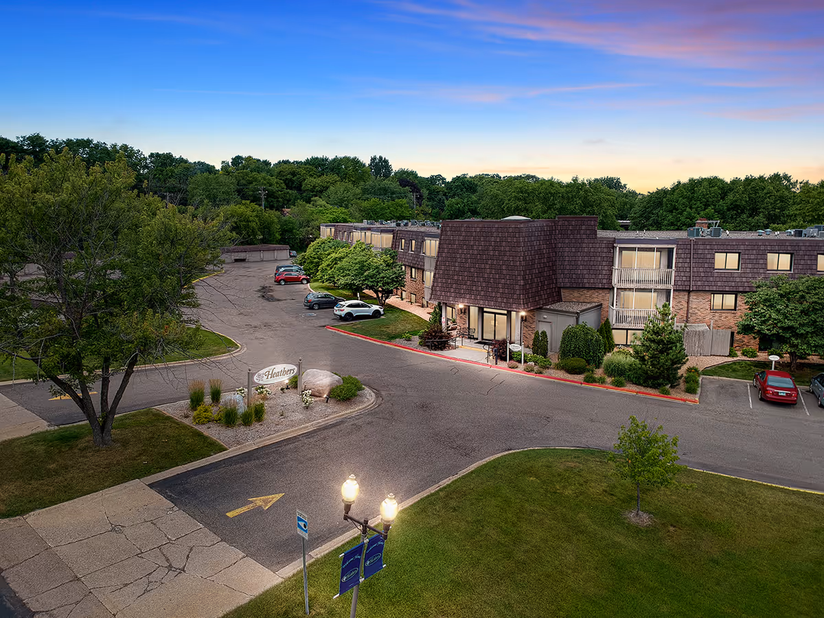 Exterior view of Heathers Estates senior living facility during dusk, showing a multi-story building with a brown shingled roof, surrounded by trees and parked cars in the driveway and parking lot.