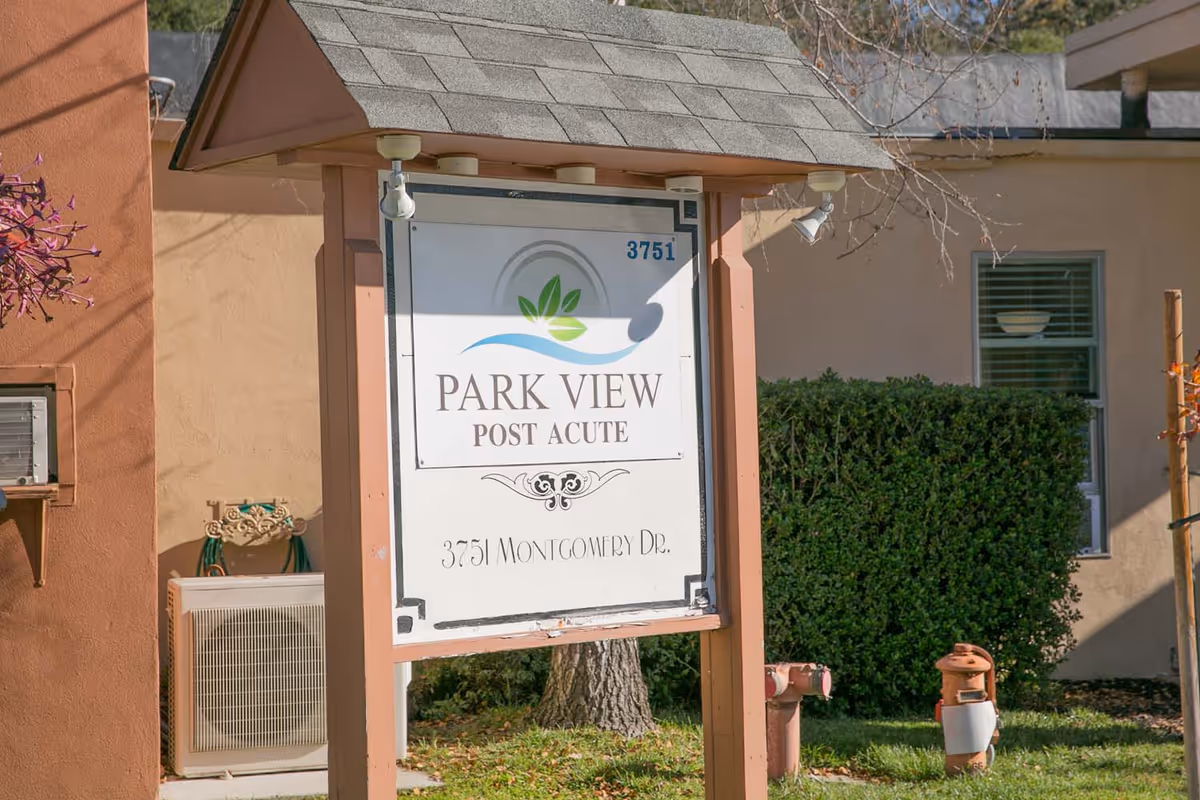 Outdoor view of a sign for Park View Post Acute located at 3751 Montgomery Dr. The sign is mounted on wooden posts with a shingled roof above it. In the background, there is a beige building with a window, some bushes, and a tree.