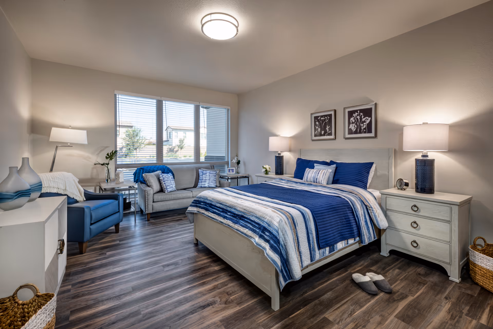 Bright furnished bedroom featuring a bed with blue-striped bedding, two nightstands with lamps, a seating area by a large window, and wood-look flooring.