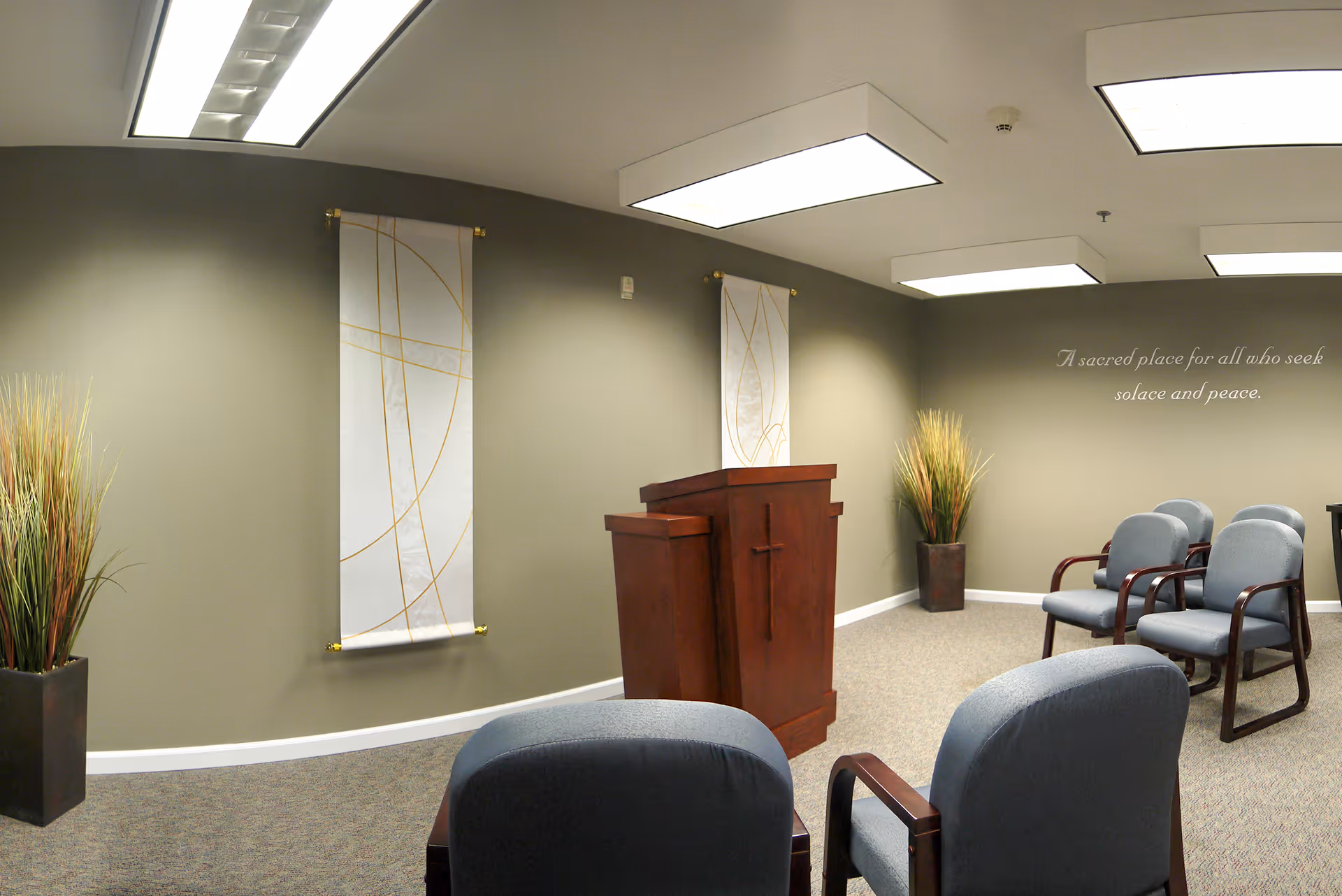 A small chapel or meditation room with a wooden podium featuring a cross, several blue cushioned chairs arranged in rows, two tall decorative plants in pots, and two white wall hangings with gold abstract lines. The walls are painted a muted green, and there is a quote on the wall that reads, 'A sacred place for all who seek solace and peace.' The room is lit by rectangular ceiling lights.
