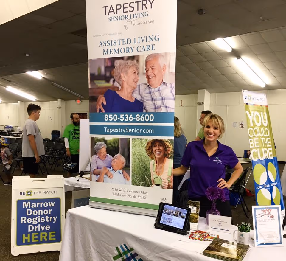 A woman in a purple Tapestry Senior Living shirt stands smiling behind a table at an indoor event. The table has brochures, a tablet, and promotional items. Behind her is a large banner for Tapestry Senior Living of Tallahassee advertising assisted living and memory care services with contact information. To the left, there is a sign for a Marrow Donor Registry Drive and people walking in the background.