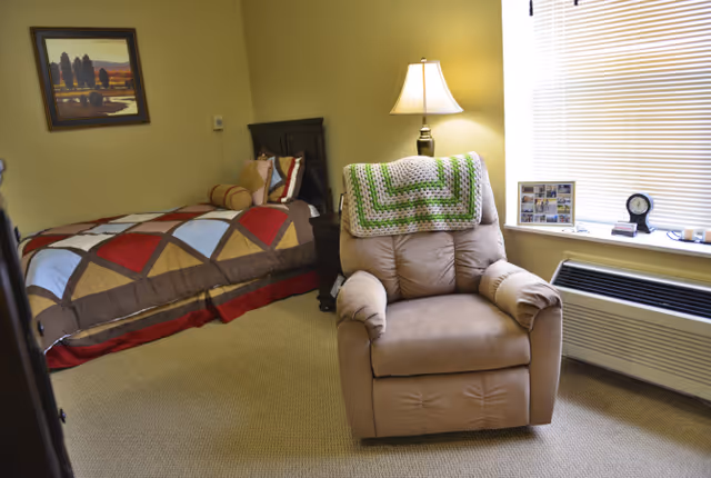A cozy bedroom with a single bed featuring a colorful quilt and pillows, a beige recliner chair with a green and white crocheted blanket draped over the back, a lit table lamp on a nightstand, a framed landscape painting on the wall, and a window with blinds partially closed above a heating/cooling unit.