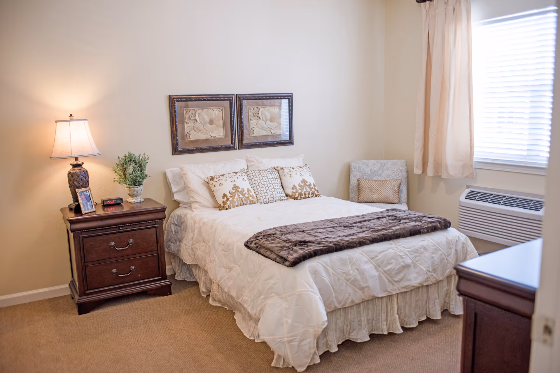 Well-lit bedroom with a neatly made bed, a bedside table with lamp and decor, an armchair, and a window air-conditioner.