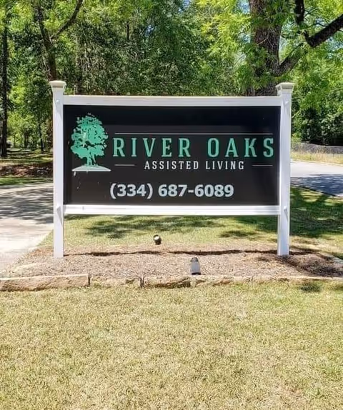 Freestanding outdoor sign reading "River Oaks Assisted Living" with a phone number on a grassy lawn and trees in the background.