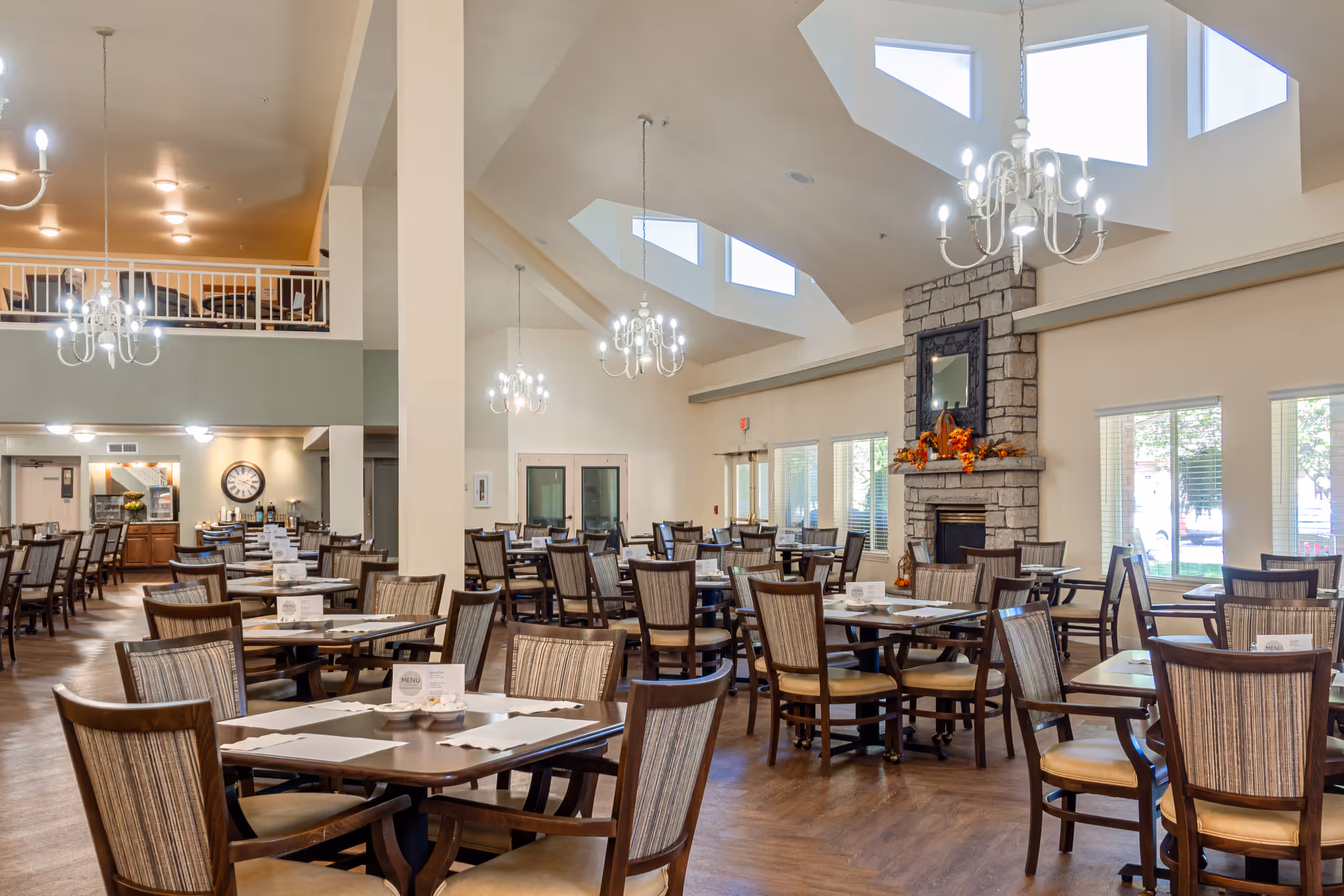 Spacious senior living dining room with multiple wooden tables and chairs, chandeliers, and a stone fireplace.
