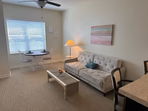 A cozy senior living room with a beige tufted sofa, a wooden coffee table, a floor lamp, and a small round dining table with two chairs near a window with blinds. A ceiling fan is visible above, and a pastel-colored abstract painting hangs on the wall.