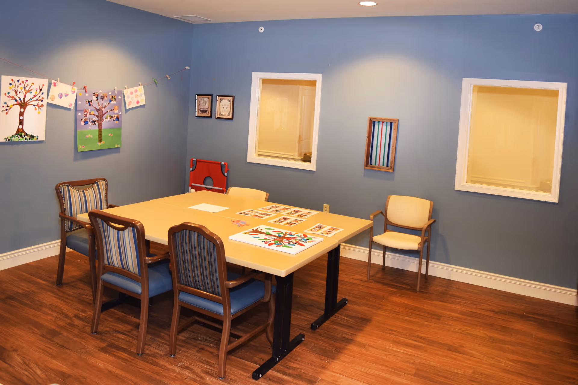 A room with blue walls and wooden flooring featuring a rectangular table surrounded by five chairs. On the table are colorful art projects and papers. The walls have framed artwork and two windows looking into another room. A string with clothespins holds additional artwork on one wall.
