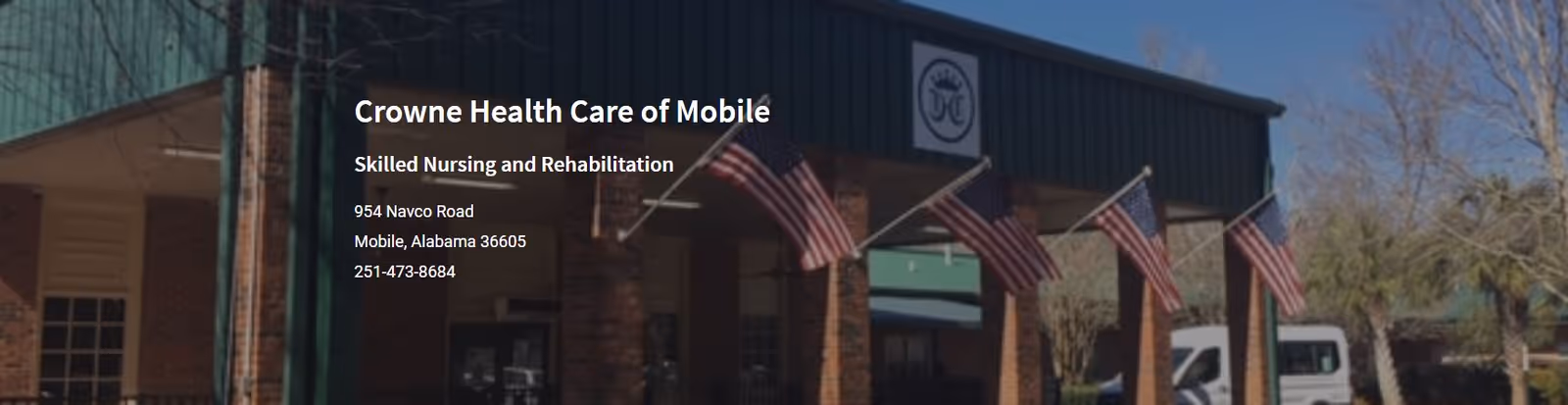 Exterior view of Crowne Health Care of Mobile building with brick pillars and multiple American flags displayed on the front. There are trees and a white vehicle visible in the background under a clear blue sky.