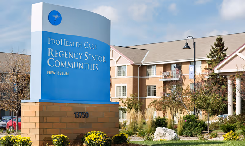 Outdoor view of ProHealth Care Regency Senior Communities New Berlin sign with a multi-story senior living building and landscaped garden in the background under a clear sky.