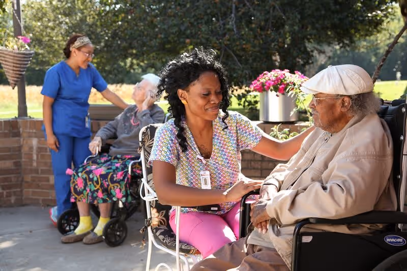 A female caregiver in colorful scrubs is sitting and gently holding the hands of an elderly man in a wheelchair outdoors. In the background, another caregiver in blue scrubs is standing and smiling while interacting with an elderly woman in a wheelchair. There are flowers and greenery around them, suggesting a garden or patio area.