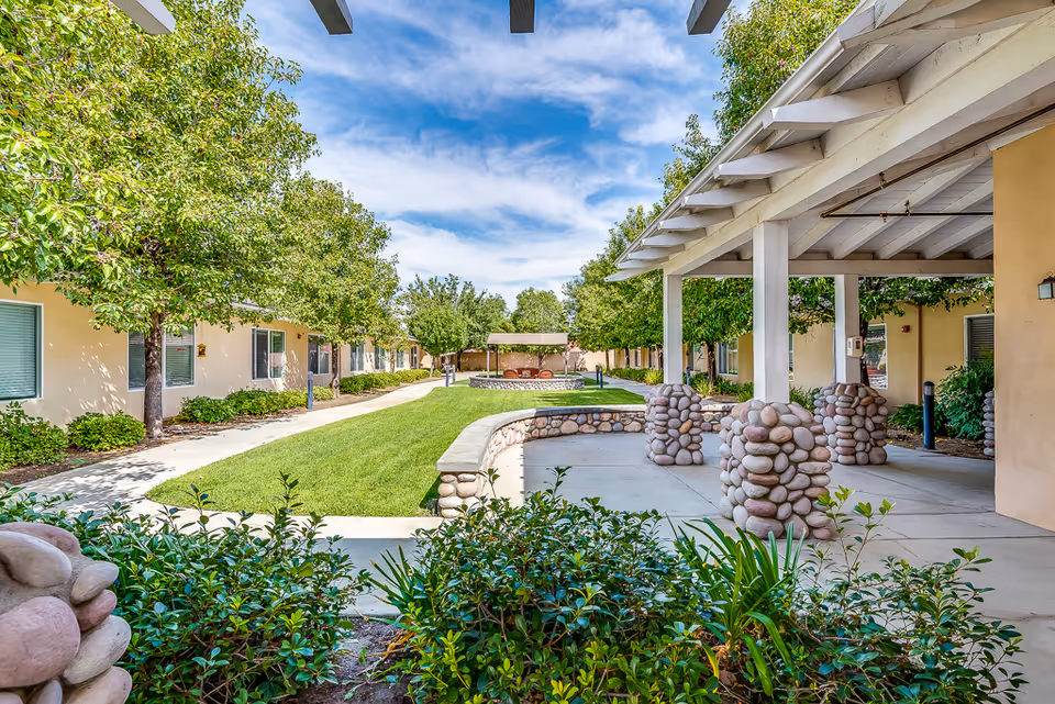 Courtyard with a covered patio supported by decorative stone columns, walkways, green lawn and trees between single-story residential buildings.