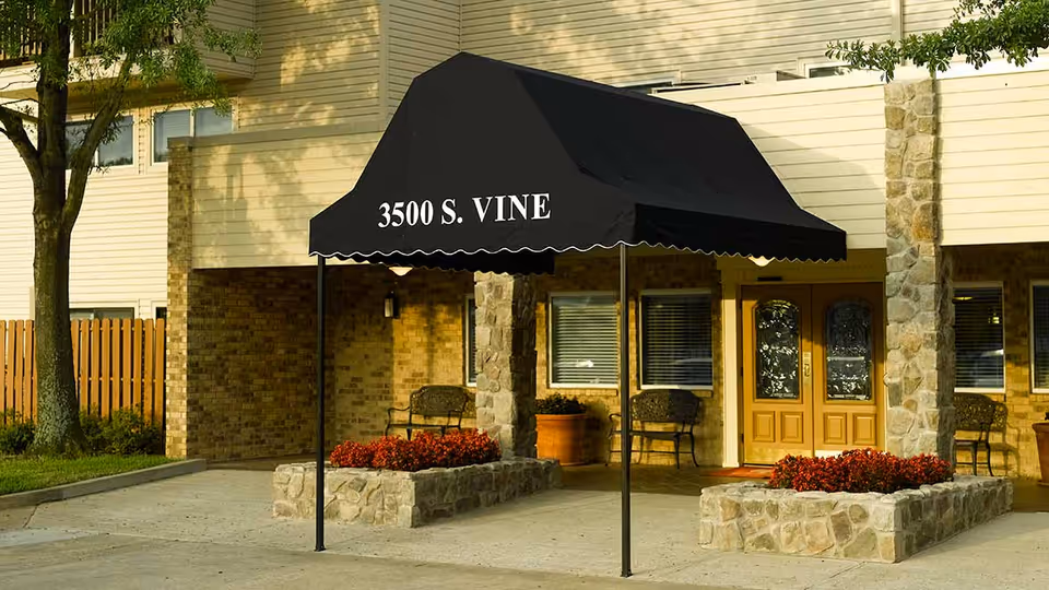Entrance of a building with a black canopy displaying the address '3500 S. VINE'. The building features stone pillars, a wooden double door with glass panels, two benches, flower beds with red flowers, and a tree on the left side.
