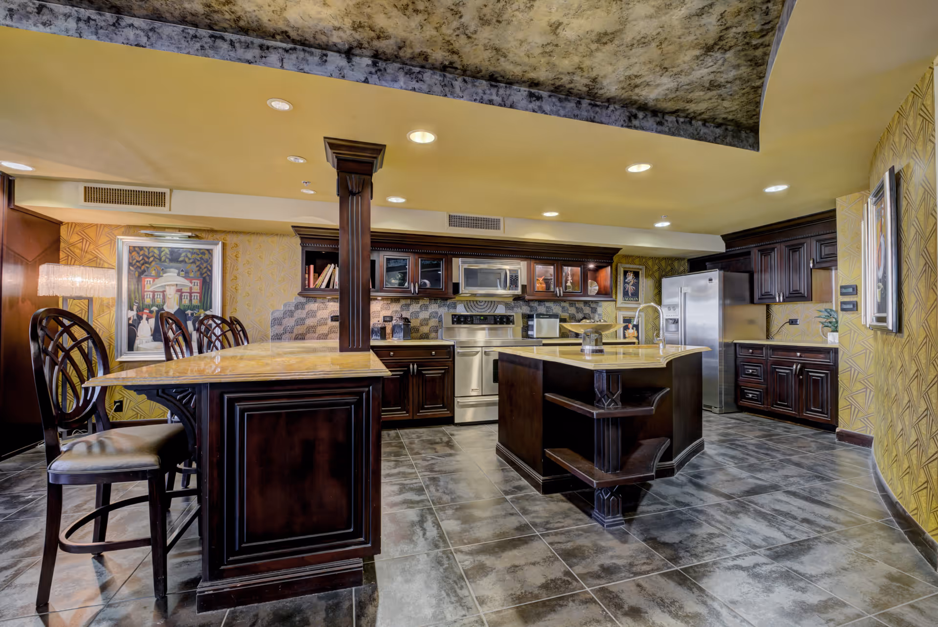 Spacious kitchen with dark wood cabinetry, a central island with a sink, stainless steel appliances including a refrigerator, stove, and microwave, and a breakfast bar with high chairs. The kitchen has tiled flooring, patterned yellow wallpaper, and recessed ceiling lights.