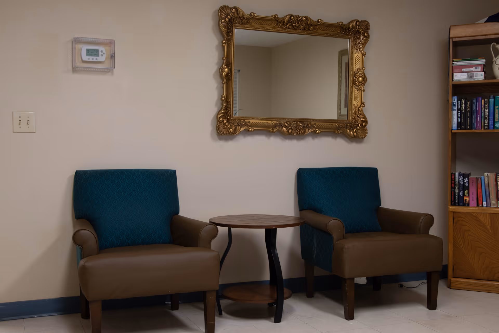 Two armchairs with brown seats and teal backs are placed on either side of a small round wooden table against a beige wall. Above the table is a large ornate gold-framed mirror. To the right, there is a wooden bookshelf filled with books and decorative items. A thermostat is mounted on the wall to the left of the chairs.