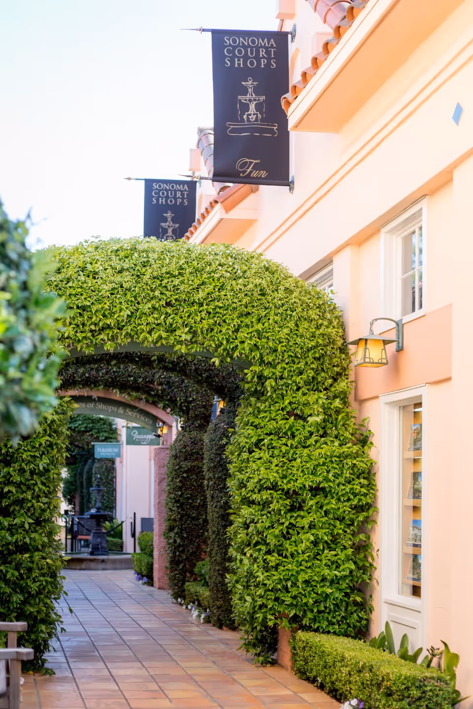 A walkway lined with neatly trimmed green archways and bushes alongside a peach-colored building with windows and outdoor wall lamps. Black banners hanging from the building read 'Sonoma Court Shops' with a fountain illustration and the word 'Fun'.