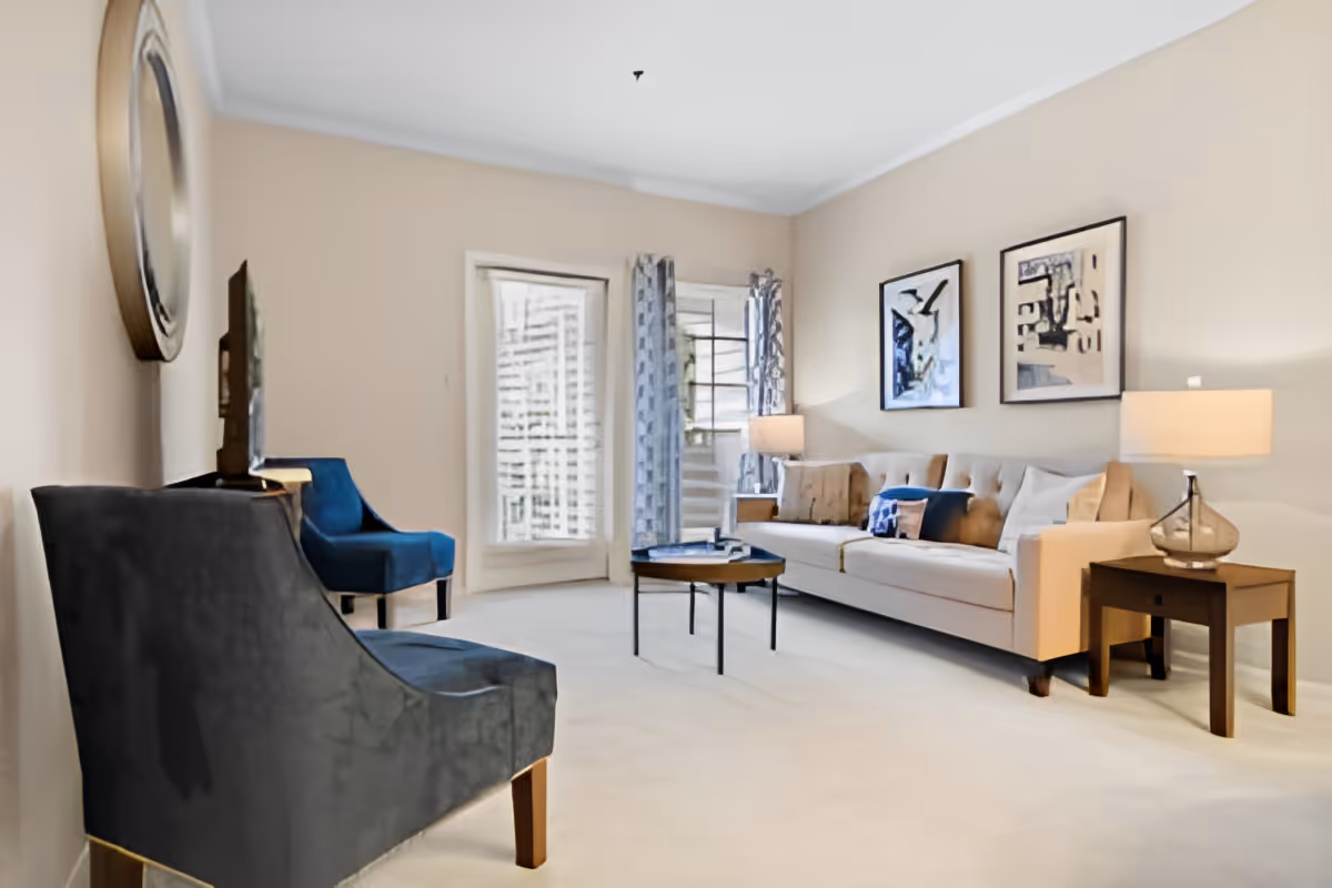 Bright living room with a beige sofa, two blue armchairs, a round coffee table, side tables with lamps, wall art, and a window with curtains.