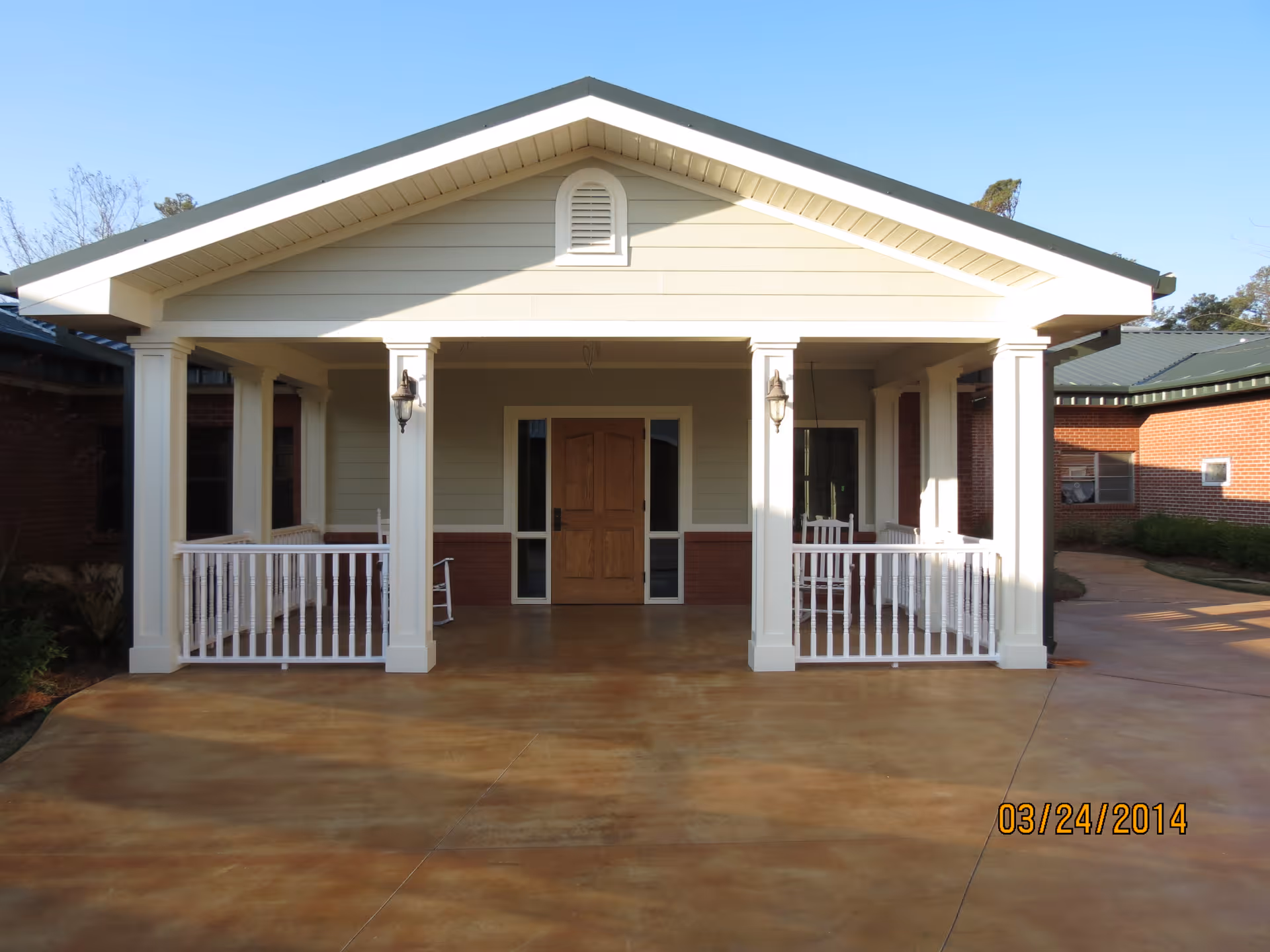 Front exterior view of a single-story building with a covered porch supported by white columns, white railings, and two rocking chairs. The building has a wooden front door and beige siding with a brick base. The sky is clear and blue.