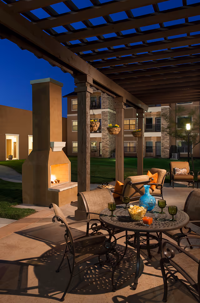 Outdoor patio area at dusk with a lit fireplace, metal table and chairs, cushioned seating with orange pillows, hanging flower pots, and a blue decorative vase on the table. The background shows a multi-story building with windows and balconies.