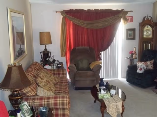 A cozy living room with a plaid sofa adorned with patterned cushions on the left, a brown recliner chair in the center, and a dark armchair with a teddy bear on the right. The room features a glass coffee table with a decorative runner and various items on it. Behind the recliner, there are red curtains with a gold swag valance covering a sliding glass door. A tall grandfather clock stands in the corner near the armchair, and a table lamp is placed on a side table next to the sofa. The walls are decorated with framed pictures and a large mirror.