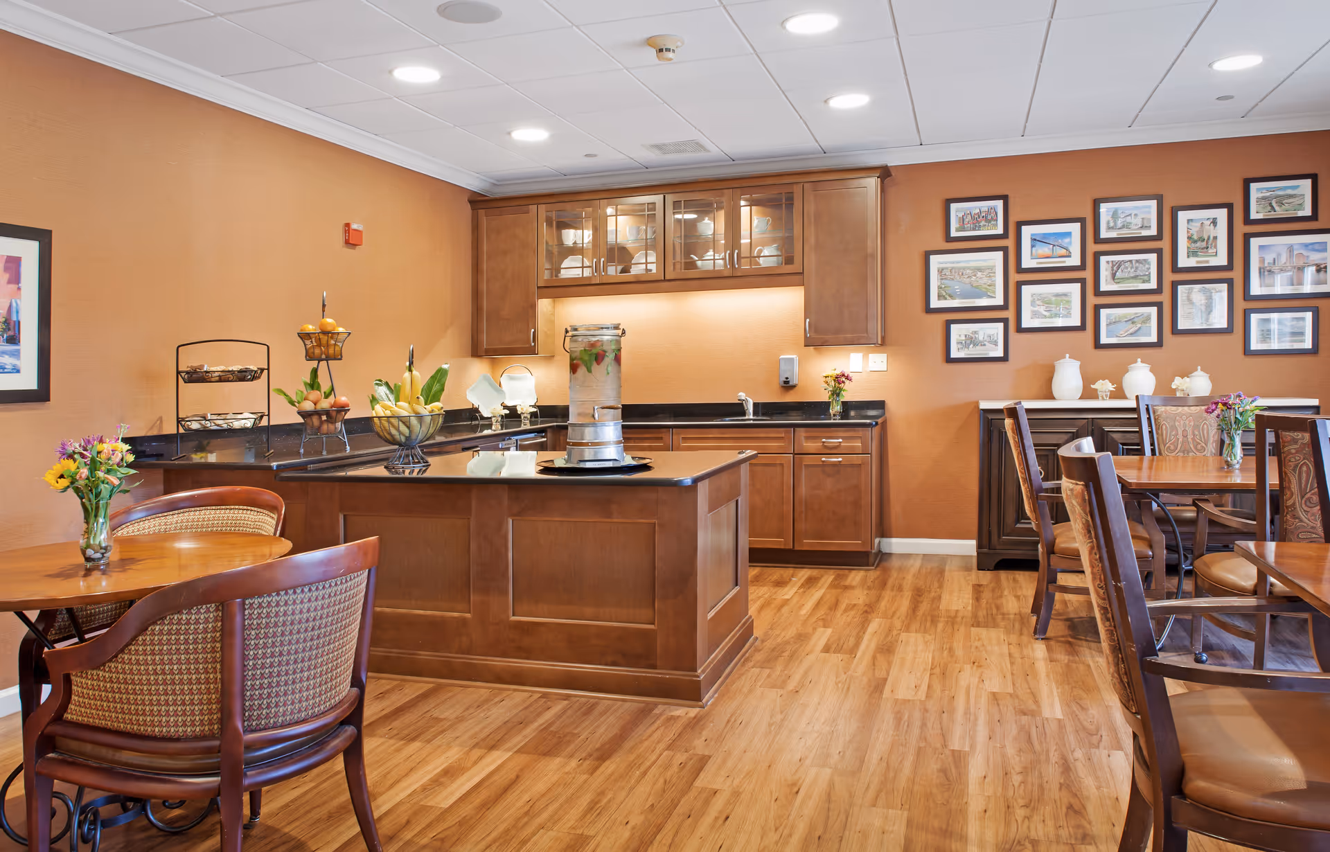 Well-lit dining area with wooden tables and chairs and a central kitchen island and cabinets against a warm-toned wall.