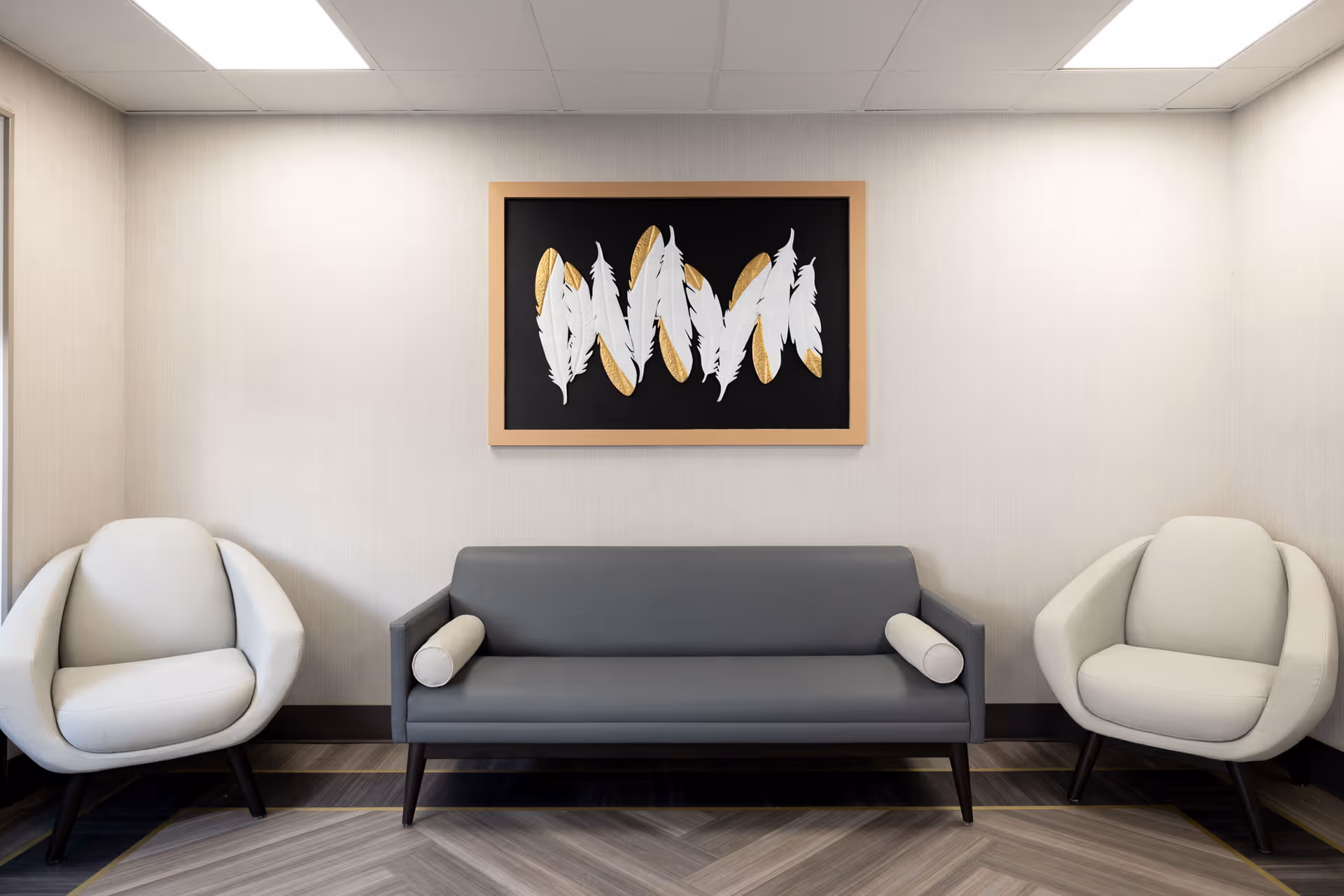 A modern seating area with a gray sofa flanked by two white armchairs. Above the sofa is a framed artwork featuring white and gold feathers on a black background. The room has light-colored walls and a patterned floor.
