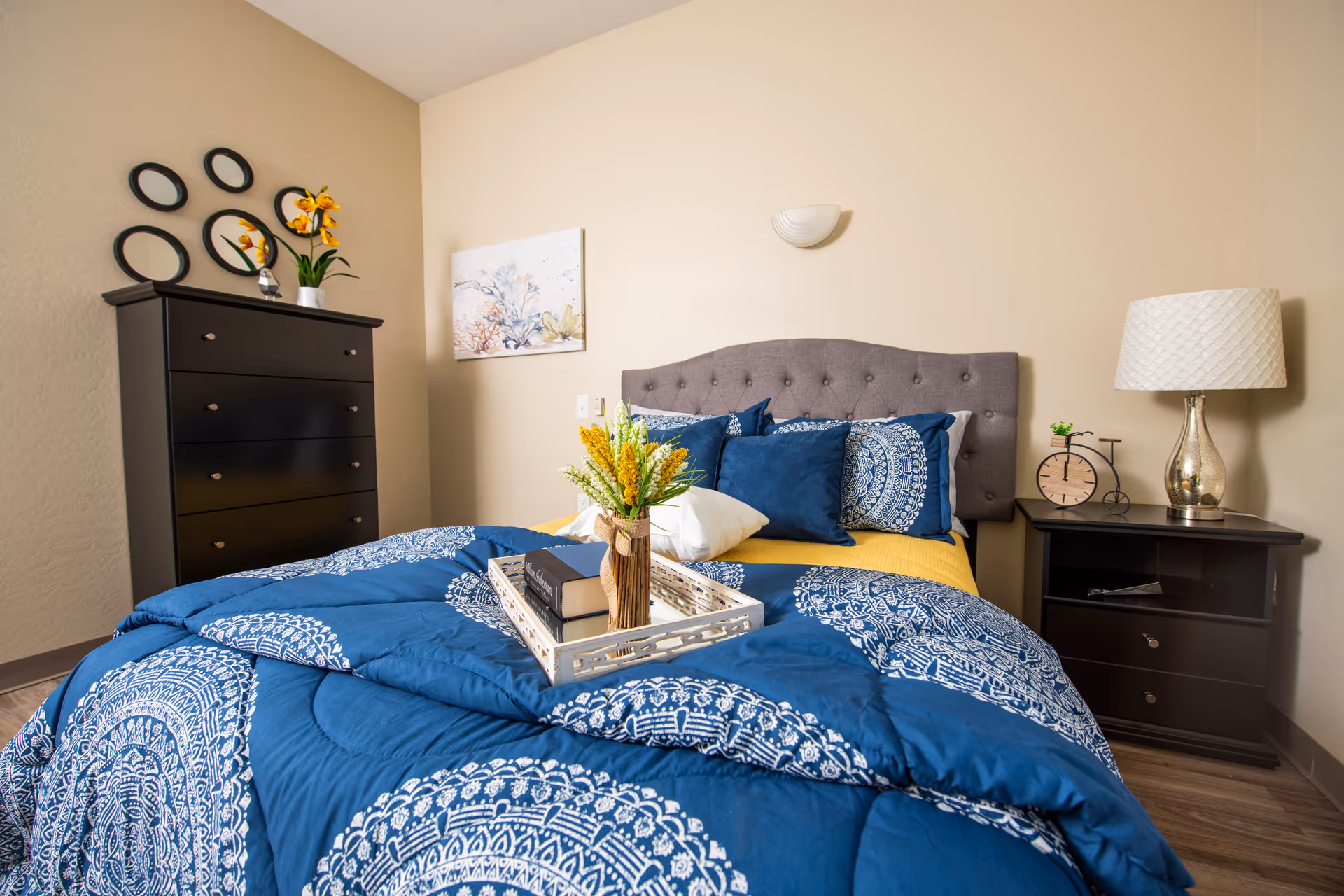 A neatly made bed with a blue and white patterned comforter and multiple pillows in a bedroom. A tray with a vase of yellow flowers and books is placed on the bed. To the right of the bed is a dark wooden nightstand with a lamp, a small clock, and a small plant. On the left side of the room is a tall dark wooden dresser with decorative round mirrors and a vase with yellow flowers on top. A framed floral artwork hangs on the wall above the bed's headboard.
