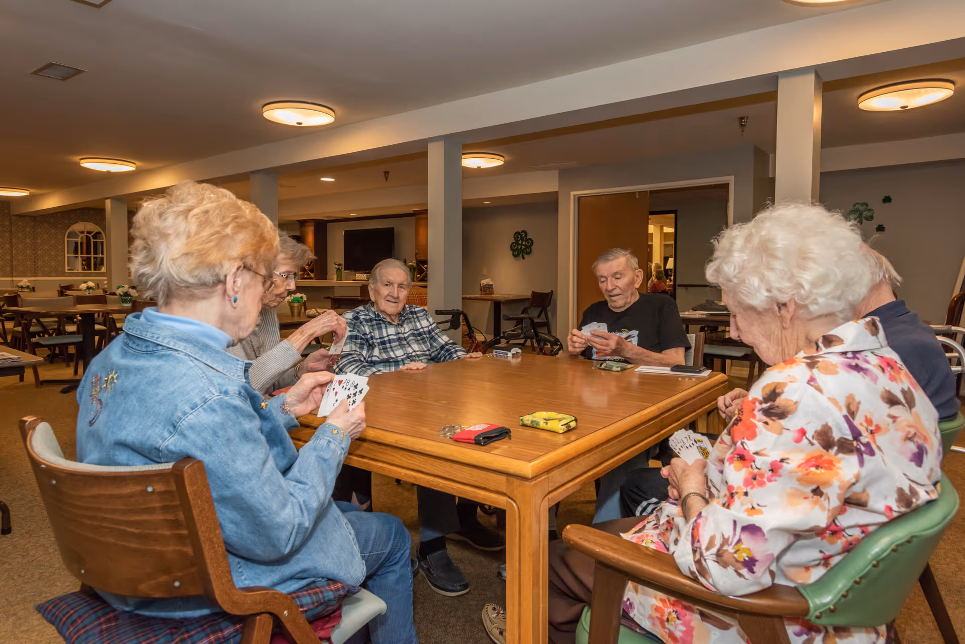A group of elderly people sitting around a wooden table playing cards in a well-lit common room with chairs and tables in the background.