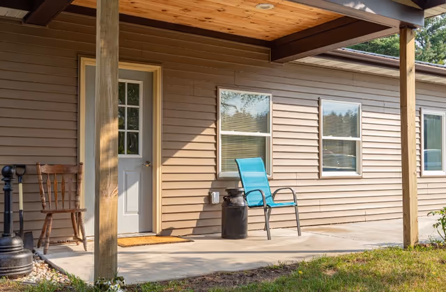 A covered outdoor patio area of a building with beige siding, featuring a white door with glass panels, two windows with blinds, a wooden chair, a blue metal chair, and a black milk can. The patio is supported by wooden posts and has a concrete floor with grass in the foreground.