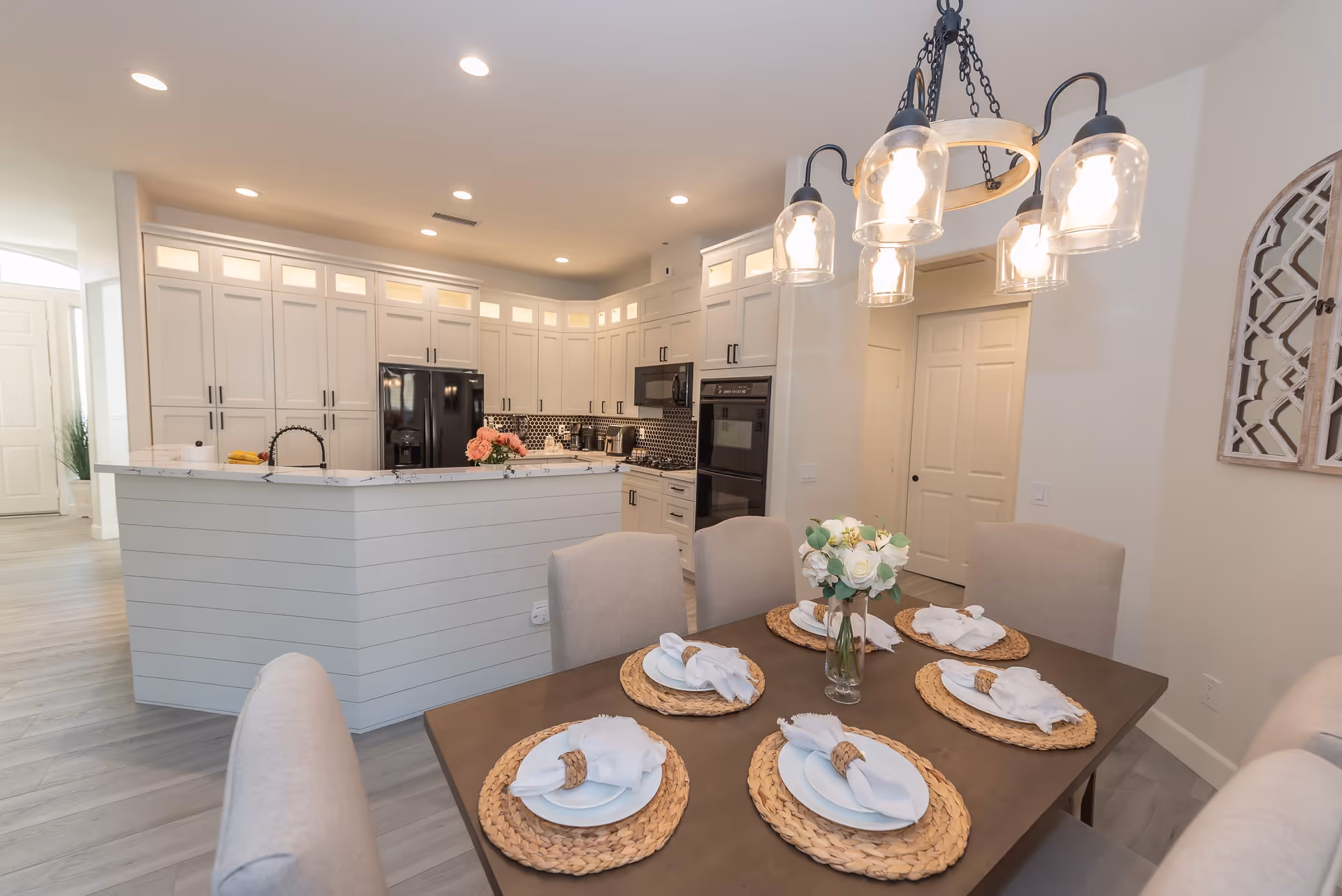 Open-concept dining area with a set table in the foreground and a modern white kitchen with an island and pendant lights in the background.