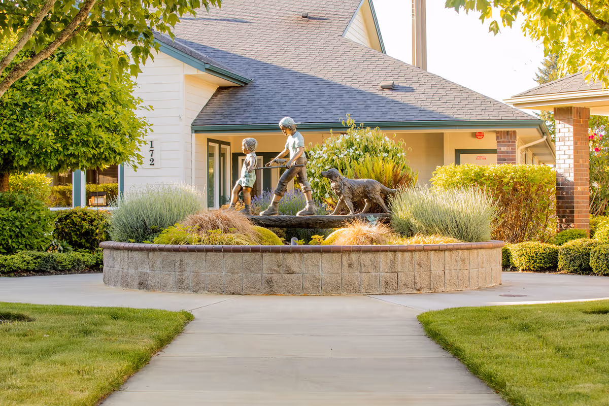 A landscaped outdoor area at Gateway Gardens featuring a circular raised stone planter with a bronze statue of two children and a dog. The background shows a building with a gray roof, beige siding, and green trim, surrounded by green bushes and trees.