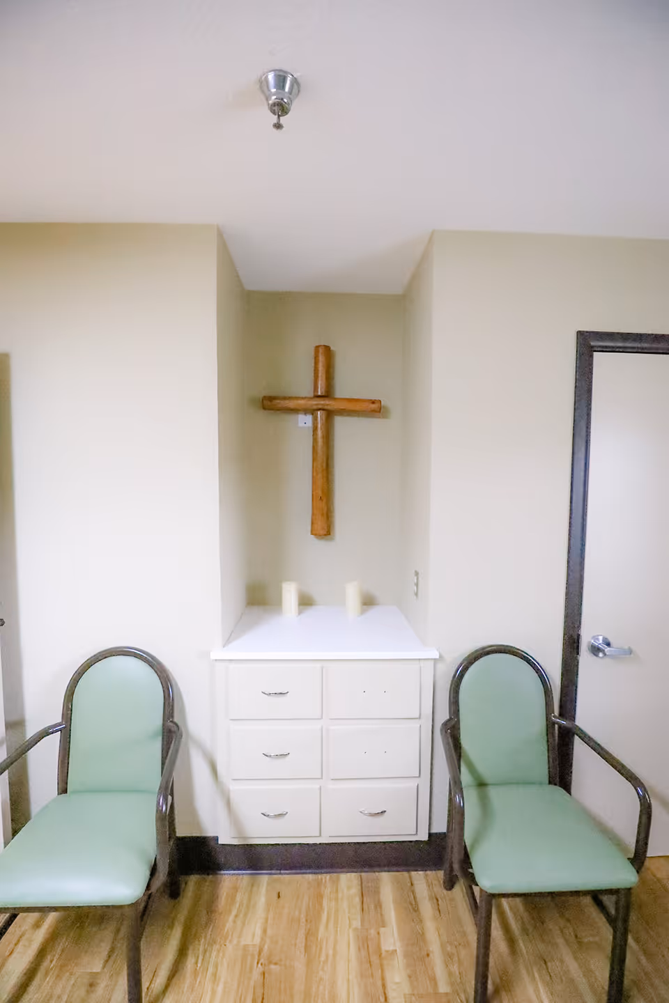 A small corner area in a room with a wooden cross mounted on the wall above a white cabinet with six drawers. Two green cushioned chairs with wooden armrests are placed on either side of the cabinet. The floor is wooden, and there is a closed door with a silver handle on the right side.