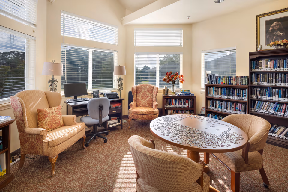 A cozy senior living community room with large windows letting in natural light. The room features a round table with a partially completed jigsaw puzzle, surrounded by four cushioned chairs. There are two armchairs with patterned upholstery, a desk with a computer and office chair, and bookshelves filled with books. A vase with flowers sits on a small bookshelf near the window.