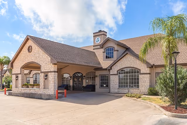 Exterior view of a single-story building with stone and stucco facade, large windows, a covered entrance with seating, and a clock tower feature on the roof. The area is landscaped with palm trees and shrubs under a clear blue sky.