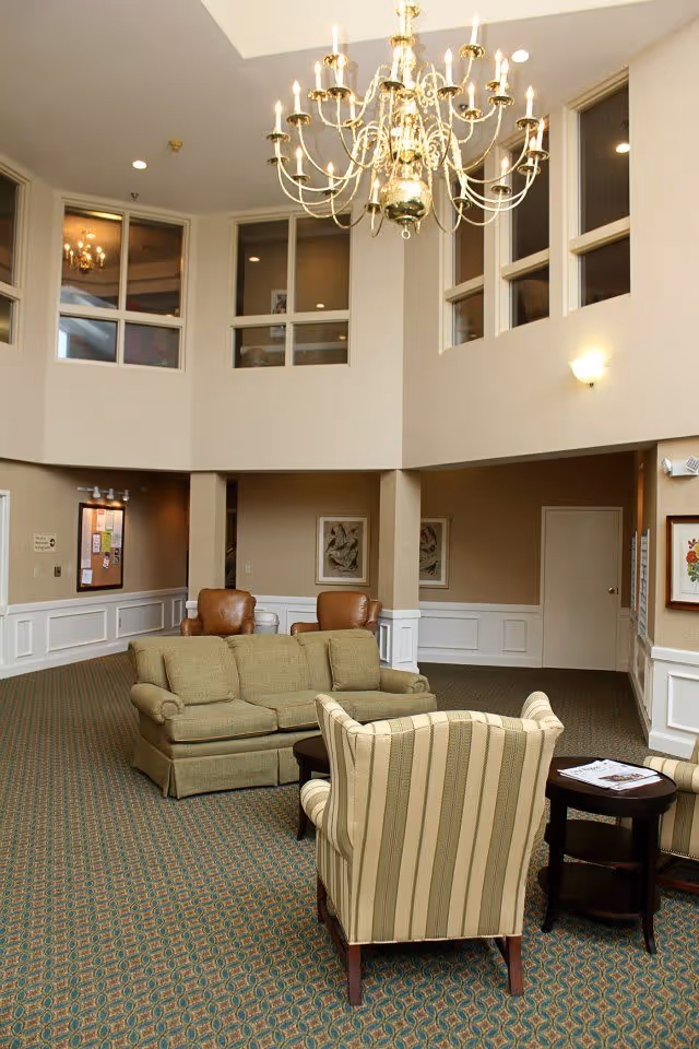 A spacious living room area with high ceilings and a large chandelier. The room features a green sofa, two brown leather chairs, and a striped armchair around a small dark wooden table. The walls are beige with white wainscoting, and there are several windows and framed artworks on the walls.