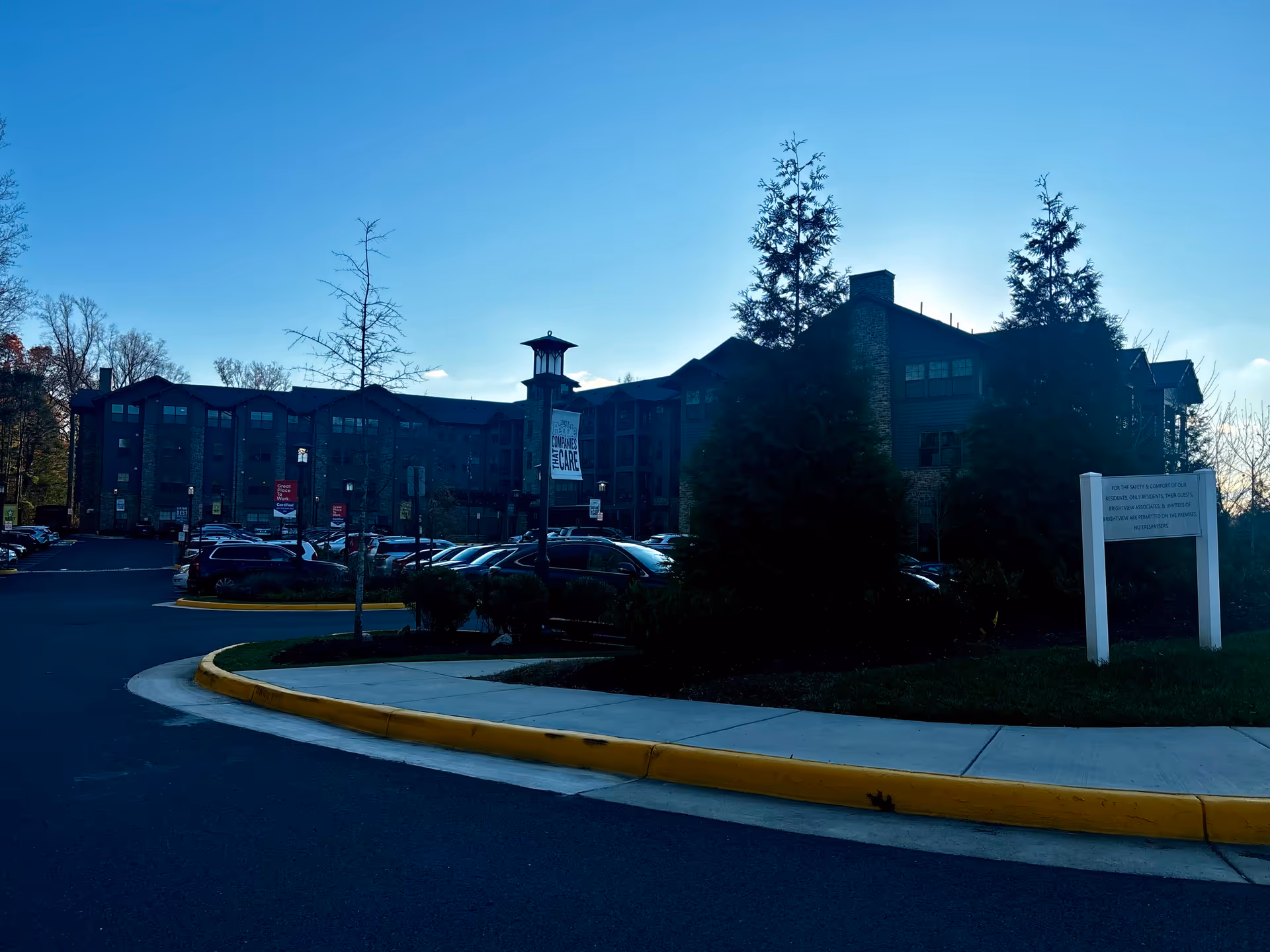 Exterior view of a senior living facility building with multiple floors, surrounded by trees and a parking lot filled with cars. There is a sidewalk with a yellow curb in the foreground and a white sign on the right side of the image. The sky is clear and blue.