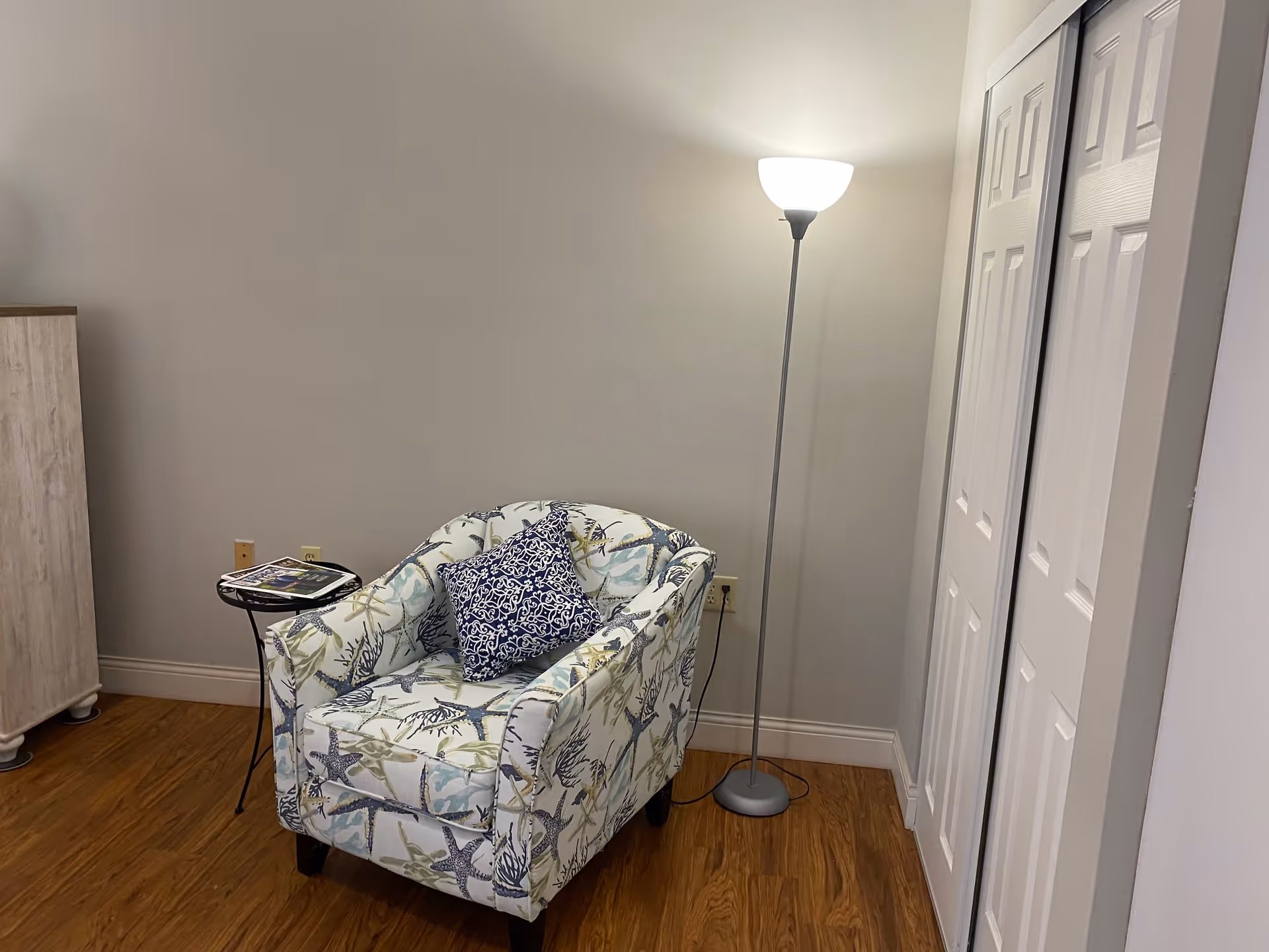 A patterned armchair with a blue decorative pillow sits next to a small side table and a tall floor lamp in a room with wood flooring and white closet doors.