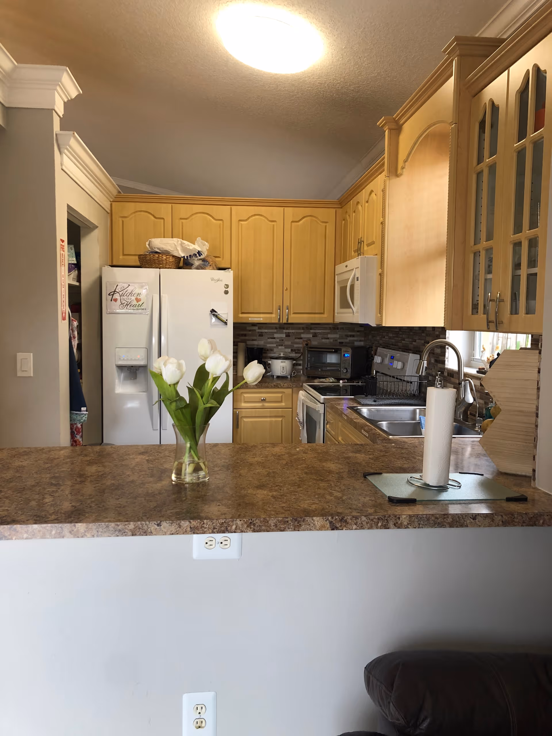 View of a residential kitchen with wooden cabinets, a refrigerator and microwave, a sink area, and a countertop island holding a vase of white tulips and a paper towel holder.