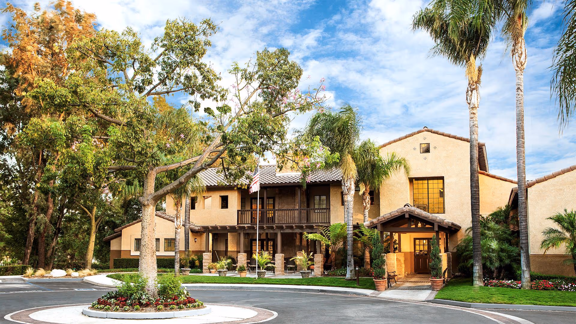 Exterior view of Atria Del Rey senior living facility showing a two-story building with a tiled roof, surrounded by palm trees and other greenery. There is a circular driveway with a landscaped island in the center featuring a tree and flowers. The sky is partly cloudy with blue patches.